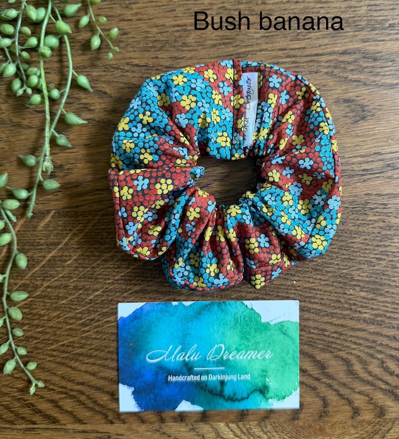 May include: A fabric hair scrunchie featuring a repeating pattern of small yellow, blue, and red flowers. A white tag is attached to the scrunchie. The text Bush banana is visible above, and a card with the text Malu Dreamer is below.