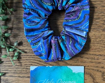 May include: A fabric scrunchie featuring a blue, purple, and black abstract swirl pattern with the text Emu Dreaming. A small white tag is attached to the side. Below is a rectangular card with a blue and green watercolor design and the text Malu Dreamer.