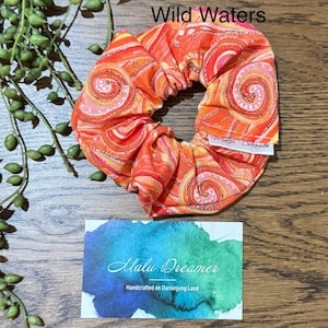 May include: A fabric hair scrunchie featuring a vibrant orange, red, and yellow spiral pattern with the text Wild Waters. A white tag reads Malu Dreamer. Below, a rectangular card displays a blue and green watercolor design with the text Malu Dreamer Handcrafted on Darkinjung Land.