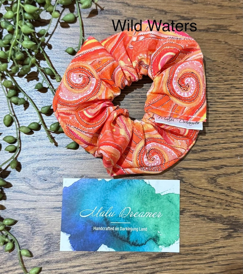 May include: A fabric hair scrunchie featuring a vibrant orange, red, and yellow spiral pattern with the text Wild Waters. A white tag reads Malu Dreamer. Below, a rectangular card displays a blue and green watercolor design with the text Malu Dreamer Handcrafted on Darkinjung Land.