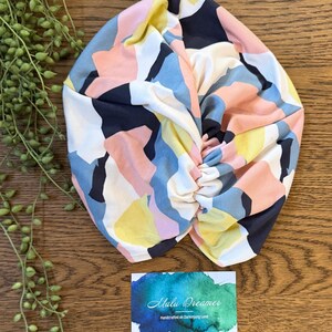 May include: A fabric head wrap featuring an abstract geometric pattern in shades of navy blue, soft pink, pale yellow, and white. The head wrap has a gathered, twisted center detail. A rectangular card with a blue and green watercolor design reads Malu Dreamer Handcrafted on Darkinjung Land.