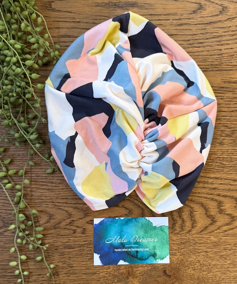 May include: A fabric head wrap featuring an abstract geometric pattern in shades of navy blue, soft pink, pale yellow, and white. The head wrap has a gathered, twisted center detail. A rectangular card with a blue and green watercolor design reads Malu Dreamer Handcrafted on Darkinjung Land.