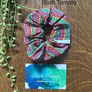 May include: A fabric hair scrunchie featuring a vibrant, multi-coloured dot pattern on a dark background. A small white tag reads Malu Dreamer. Above the scrunchie, the text reads Bush Tomato. Below, a card displays the text Malu Dreamer Handcrafted on Darkinjung Land.