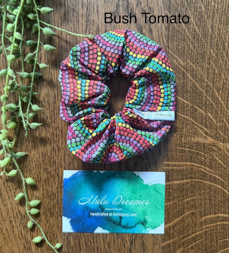 May include: A fabric hair scrunchie featuring a vibrant, multi-coloured dot pattern on a dark background. A small white tag reads Malu Dreamer. Above the scrunchie, the text reads Bush Tomato. Below, a card displays the text Malu Dreamer Handcrafted on Darkinjung Land.