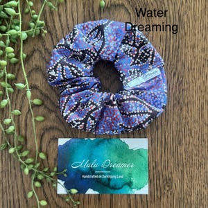 May include: A fabric hair scrunchie featuring a pattern of blue, red, and black dots. A small white tag on the scrunchie reads Malu Dreamer. The text Water Dreaming is visible above the scrunchie, and a card below reads Malu Dreamer Handcrafted on Darkinjung Land.