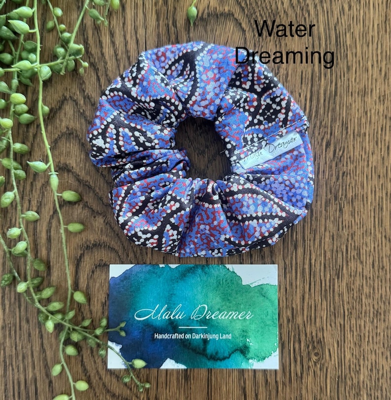 May include: A fabric hair scrunchie featuring a pattern of blue, red, and black dots. A small white tag on the scrunchie reads Malu Dreamer. The text Water Dreaming is visible above the scrunchie, and a card below reads Malu Dreamer Handcrafted on Darkinjung Land.