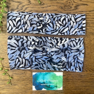 May include: Two fabric headbands with a twisted knot design. The headbands have a repeating pattern of blue, white, and grey brushstrokes on a black background. A small card with the text "Malu Dreamer" is visible.