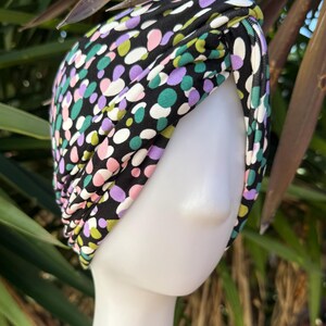 May include: A black turban featuring a pattern of multi-coloured polka dots in pink, green, purple, and white. The turban is displayed on a white mannequin head, with green foliage in the background.