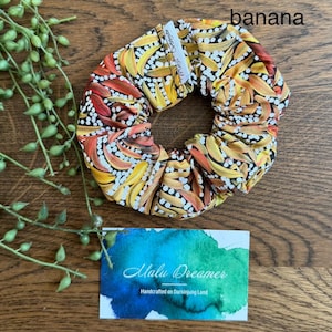 May include: A fabric hair scrunchie featuring a repeating pattern of yellow, orange, and red banana shapes on a black background with white dots. A small white tag reads Malu Dreamer. A card below displays the text Malu Dreamer Handcrafted on Darkinjung Land.
