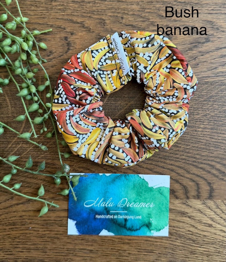 May include: A fabric hair scrunchie featuring a repeating pattern of yellow, orange, and red banana shapes on a black background with white dots. A small white tag reads Malu Dreamer. A card below displays the text Malu Dreamer Handcrafted on Darkinjung Land.