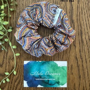 May include: A fabric hair scrunchie featuring a repeating pattern of orange, blue, and white dots arranged in swirling lines. A small white tag labeled Malu Dreamer is attached. The word Mina is printed above the scrunchie, and a card below reads Malu Dreamer Handcrafted on Darkinjung Land.