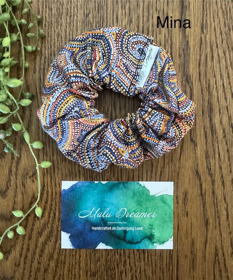 May include: A fabric hair scrunchie featuring a repeating pattern of orange, blue, and white dots arranged in swirling lines. A small white tag labeled Malu Dreamer is attached. The word Mina is printed above the scrunchie, and a card below reads Malu Dreamer Handcrafted on Darkinjung Land.