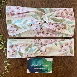 May include: Two floral headbands with a cream background and pink flowers, green leaves, and blue accents. The headbands are twisted at the top. A business card with the text "Malu Dreamer" is at the bottom.