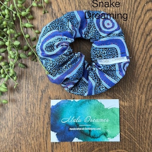 May include: A blue and black fabric scrunchie with a patterned design featuring swirling lines and dots, labeled Snake Dreaming. A white tag reads Malu Dreamer. A rectangular card features a blue and green watercolor wash and the text Malu Dreamer Handcrafted on Darkinjung Land.