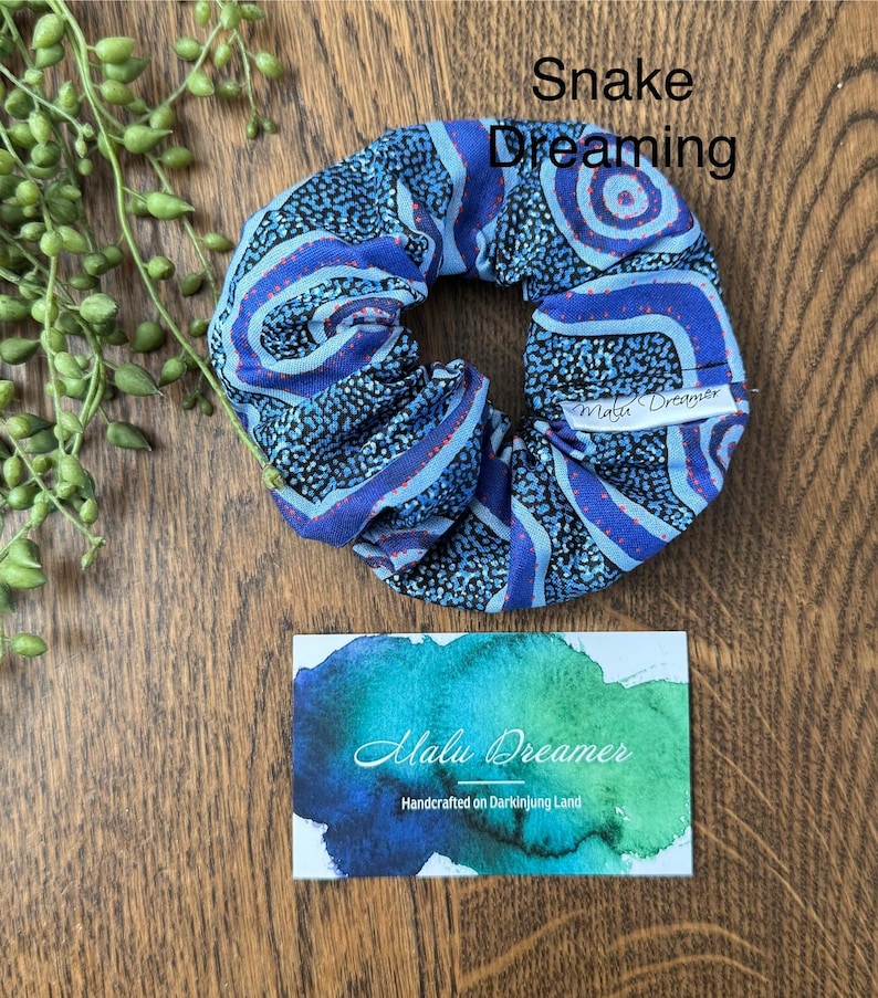 May include: A blue and black fabric scrunchie with a patterned design featuring swirling lines and dots, labeled Snake Dreaming. A white tag reads Malu Dreamer. A rectangular card features a blue and green watercolor wash and the text Malu Dreamer Handcrafted on Darkinjung Land.