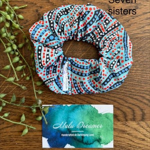 May include: A fabric hair scrunchie featuring a light blue background with a repeating pattern of black curved lines and red and blue dots. A small white tag reads Malu Dreamer. Text in the image reads Seven sisters.