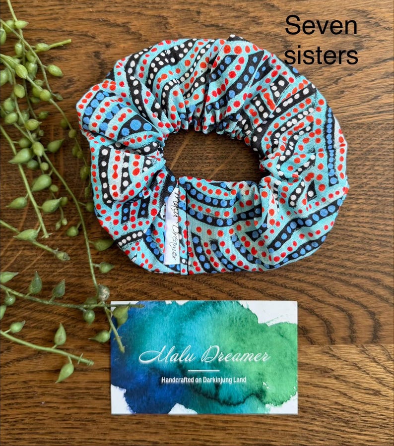 May include: A fabric hair scrunchie featuring a light blue background with a repeating pattern of black curved lines and red and blue dots. A small white tag reads Malu Dreamer. Text in the image reads Seven sisters.