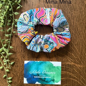 May include: A fabric hair scrunchie featuring a vibrant, multi-colored abstract pattern of pink, orange, light blue, and black swirls. A small white tag labeled Mina Mina is attached. A rectangular card displays the text Malu Dreamer Handcrafted on Darkinjung Land.