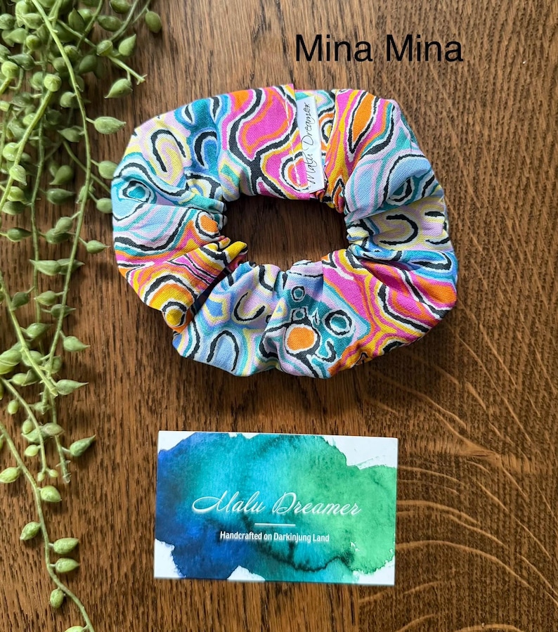 May include: A fabric hair scrunchie featuring a vibrant, multi-colored abstract pattern of pink, orange, light blue, and black swirls. A small white tag labeled Mina Mina is attached. A rectangular card displays the text Malu Dreamer Handcrafted on Darkinjung Land.