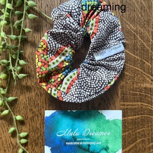 May include: A fabric scrunchie featuring a dot-patterned design in black, white, orange, yellow, green, and blue. A white tag reads Malu Dreamer. The text Goanna dreaming appears above. A rectangular card displays the text Malu Dreamer Handcrafted on Darkinjung Land.