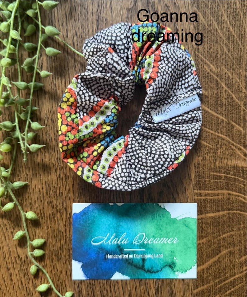 May include: A fabric scrunchie featuring a dot-patterned design in black, white, orange, yellow, green, and blue. A white tag reads Malu Dreamer. The text Goanna dreaming appears above. A rectangular card displays the text Malu Dreamer Handcrafted on Darkinjung Land.
