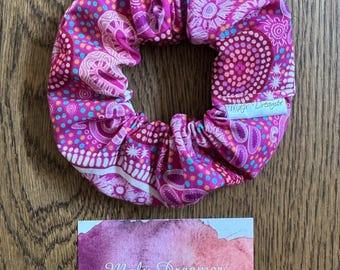May include: A pink fabric hair scrunchie featuring a multi-coloured dot and floral pattern with a white tag labelled Malu Dreamer. Below is a rectangular card with a purple watercolour background and the text Malu Dreamer Handcrafted gifts.