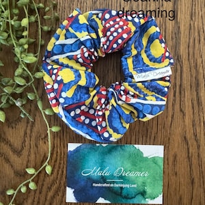 May include: A fabric hair scrunchie featuring a vibrant pattern of blue, yellow, and red shapes with white dots. The text Goanna dreaming is visible above the scrunchie, and a white tag reads Malu Dreamer. A card below displays the text Malu Dreamer Handcrafted on Darkinjung Land.