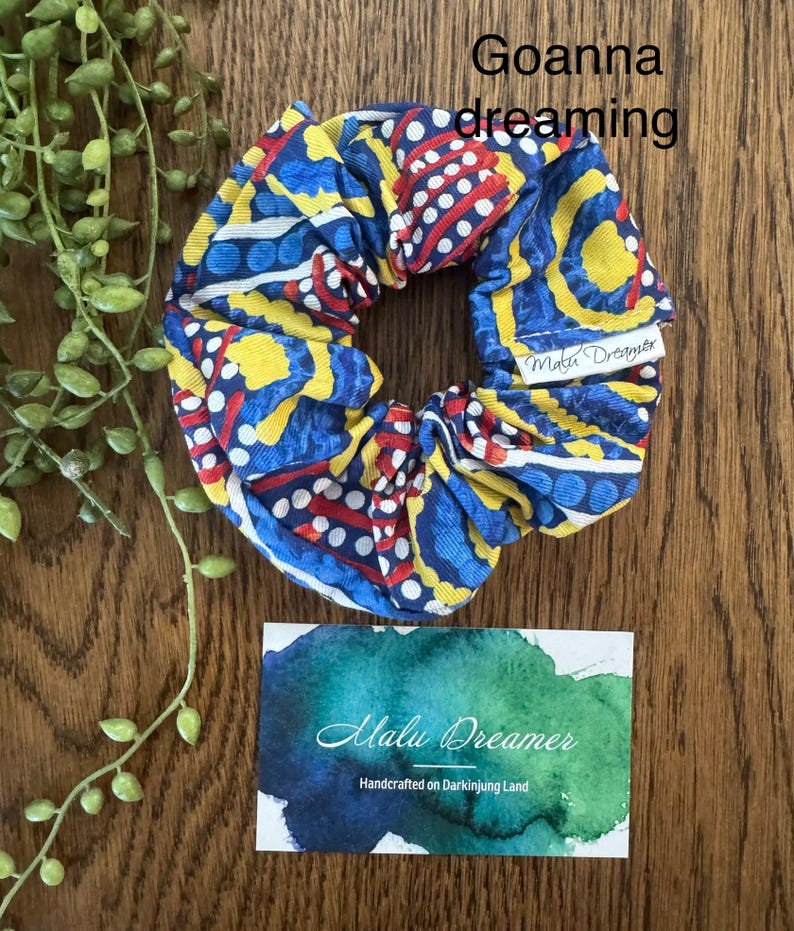 May include: A fabric hair scrunchie featuring a vibrant pattern of blue, yellow, and red shapes with white dots. The text Goanna dreaming is visible above the scrunchie, and a white tag reads Malu Dreamer. A card below displays the text Malu Dreamer Handcrafted on Darkinjung Land.