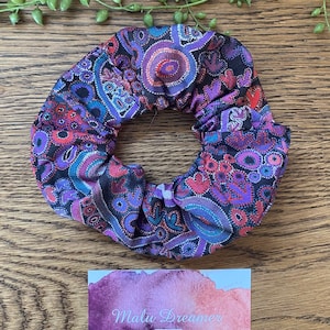May include: A fabric hair scrunchie featuring a black base with a vibrant, multi-coloured pattern of purple, blue, and red circular and organic shapes. Below the scrunchie is a rectangular card with a watercolour design and the text Malu Dreamer.