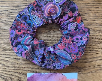 May include: A fabric hair scrunchie featuring a black base with a vibrant, multi-colored pattern of purple, blue, and red circular and organic shapes. Below the scrunchie is a rectangular card with a watercolor design and the text Malu Dreamer.
