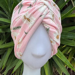 May include: A pink turban featuring a white daisy floral design. The turban is knotted at the top and displayed on a white mannequin head. Green leaves are visible in the background.