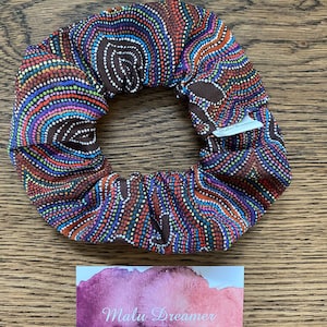 May include: A fabric hair scrunchie featuring a dark brown base with a vibrant dot art pattern in shades of orange, blue, purple, and green. A small white tag reads Malu Dreamer. Below, a rectangular card displays the text Malu Dreamer.