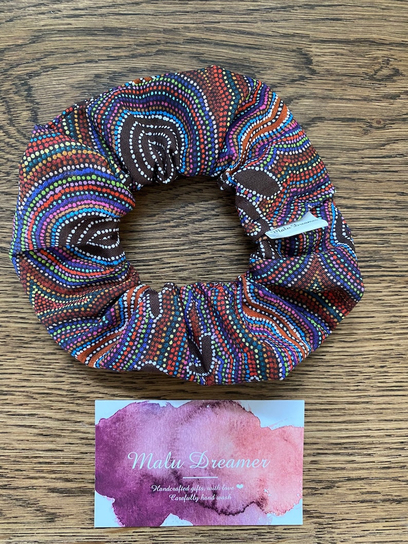 May include: A fabric hair scrunchie featuring a dark brown base with a vibrant dot art pattern in shades of orange, blue, purple, and green. A small white tag reads Malu Dreamer. Below, a rectangular card displays the text Malu Dreamer.
