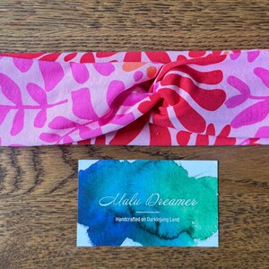 May include: A pink and red floral print headband with a twisted knot design. The headband is made of soft fabric and features a vibrant pattern of leaves and flowers. A small card with the text "Malu Dreamer" is also visible.
