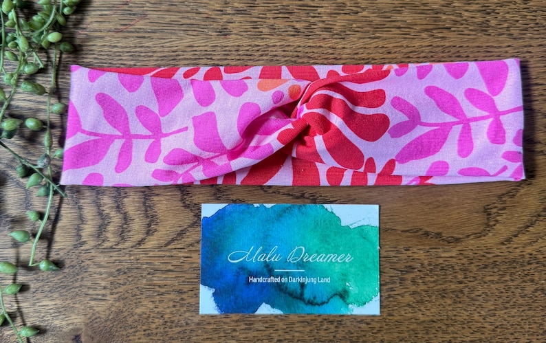 May include: A pink and red floral print headband with a twisted knot design. The headband is made of soft fabric and features a vibrant pattern of leaves and flowers. A small card with the text "Malu Dreamer" is also visible.