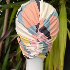 May include: A fabric turban head covering featuring a multi-colored abstract pattern with shapes in shades of peach, light blue, yellow, and dark gray. The headpiece has a gathered, ruched design running vertically down the center back.