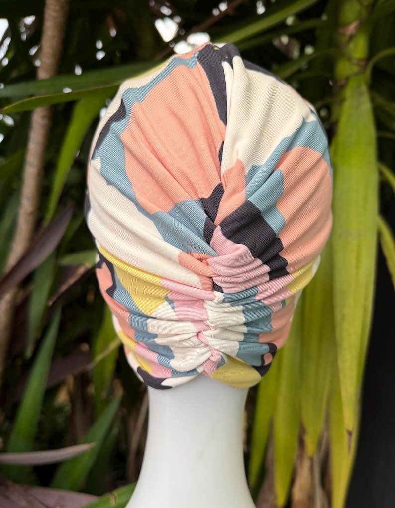 May include: A fabric turban head covering featuring a multi-colored abstract pattern with shapes in shades of peach, light blue, yellow, and dark gray. The headpiece has a gathered, ruched design running vertically down the center back.