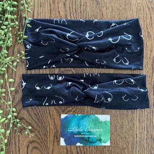 May include: Two black headbands with a white line drawing pattern of breasts. The headbands are twisted at the top. A small watercolour business card is at the bottom.