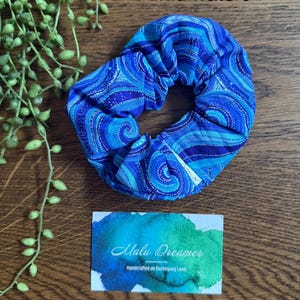 May include: A fabric hair scrunchie featuring a blue and turquoise swirling pattern with the text Wild waters. A small white tag is attached to the scrunchie. A rectangular card displays the text Malu Dreamer Handcrafted on Darkinjung Land.