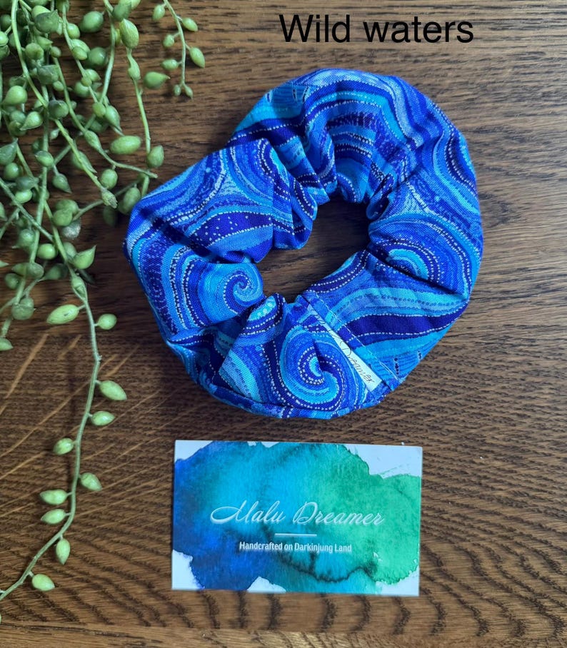 May include: A fabric hair scrunchie featuring a blue and turquoise swirling pattern with the text Wild waters. A small white tag is attached to the scrunchie. A rectangular card displays the text Malu Dreamer Handcrafted on Darkinjung Land.