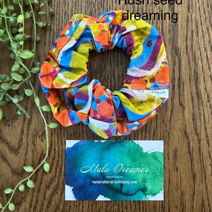 May include: A fabric scrunchie featuring a vibrant abstract pattern of orange, yellow, blue, and purple circles and waves. A small white tag reads Malu Dreamer. Text above reads Rush seed dreaming and a card below reads Malu Dreamer Handcrafted on Darkinjung Land.