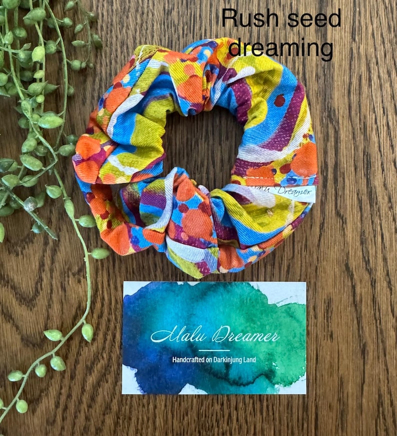 May include: A fabric scrunchie featuring a vibrant abstract pattern of orange, yellow, blue, and purple circles and waves. A small white tag reads Malu Dreamer. Text above reads Rush seed dreaming and a card below reads Malu Dreamer Handcrafted on Darkinjung Land.