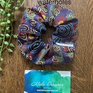 May include: A fabric hair scrunchie featuring a dark purple, blue, and orange dot-patterned design with U-shaped motifs. A small white tag reads Malu Dreamer. Text above reads Women's gathering waterholes. A card below reads Malu Dreamer Handcrafted on Darkinjung Land.