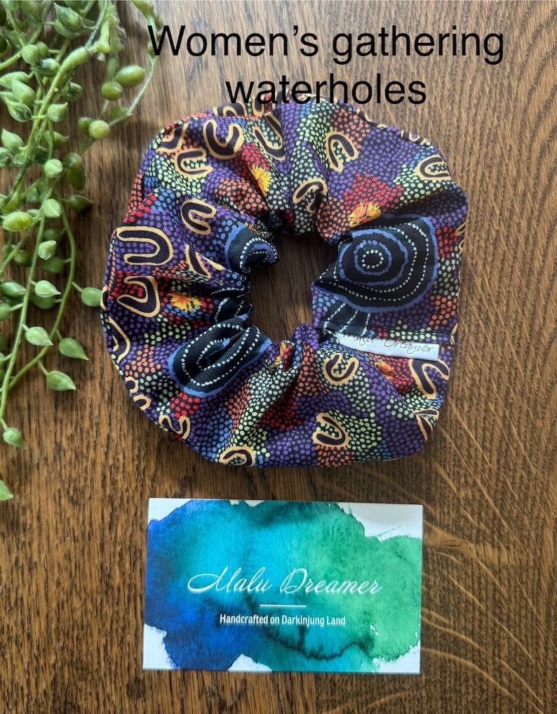 May include: A fabric hair scrunchie featuring a dark purple, blue, and orange dot-patterned design with U-shaped motifs. A small white tag reads Malu Dreamer. Text above reads Women's gathering waterholes. A card below reads Malu Dreamer Handcrafted on Darkinjung Land.