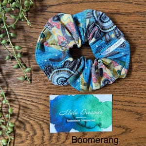 May include: A fabric hair scrunchie featuring a multi-coloured pattern of blue, yellow, and red lines and circular motifs. A small white tag reads Malu Dreamer. Below, a rectangular card displays a blue and green watercolour wash with the text Malu Dreamer Handcrafted on Darkinjung Land and the word Boomerang.