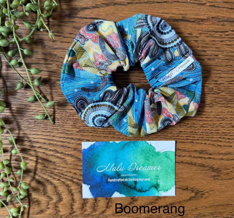 May include: A fabric hair scrunchie featuring a multi-coloured pattern of blue, yellow, and red lines and circular motifs. A small white tag reads Malu Dreamer. Below, a rectangular card displays a blue and green watercolour wash with the text Malu Dreamer Handcrafted on Darkinjung Land and the word Boomerang.