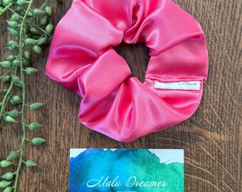 May include: A bright pink satin hair scrunchie with a small white fabric tag labeled Malu Dreamer. Below the scrunchie is a rectangular card featuring a blue and green watercolor background with the text Malu Dreamer Handcrafted on Darkinjung Land.