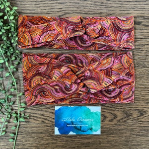 May include: Two red, orange and pink patterned headbands with a knotted design. The headbands are made of fabric with a repeating pattern of curved lines and dots. A business card with the text "Malu Dreamer" and "Handcrafted on Darug Land" is in the bottom right corner of the image.