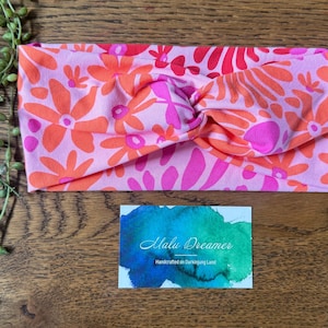 May include: A pink floral headband featuring orange and fuchsia flowers. The headband has a twisted knot design. A business card with the text "Malu Dreamer" is below the headband.