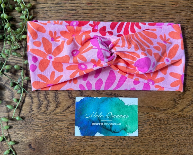 May include: A pink floral headband featuring orange and fuchsia flowers. The headband has a twisted knot design. A business card with the text "Malu Dreamer" is below the headband.